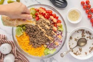Shredded cheddar cheese and pickles layered on top of seasoned beef and chopped lettuce in a glass bowl, topped with salad dressing.