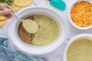 Green chile cream sauce being spooned over seasoned chicken breasts in a white crockpot, with tortillas and shredded cheese nearby.