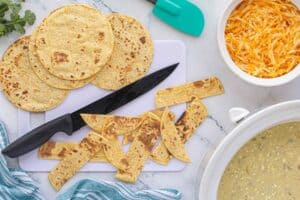 Toasted corn tortillas cut into strips on a white cutting board with a knife, next to a bowl of shredded cheese and a crockpot filled with green chile sauce.