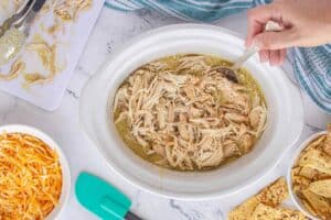 Shredded chicken being stirred into green chile sauce in a white crockpot, surrounded by shredded cheese and toasted tortillas.