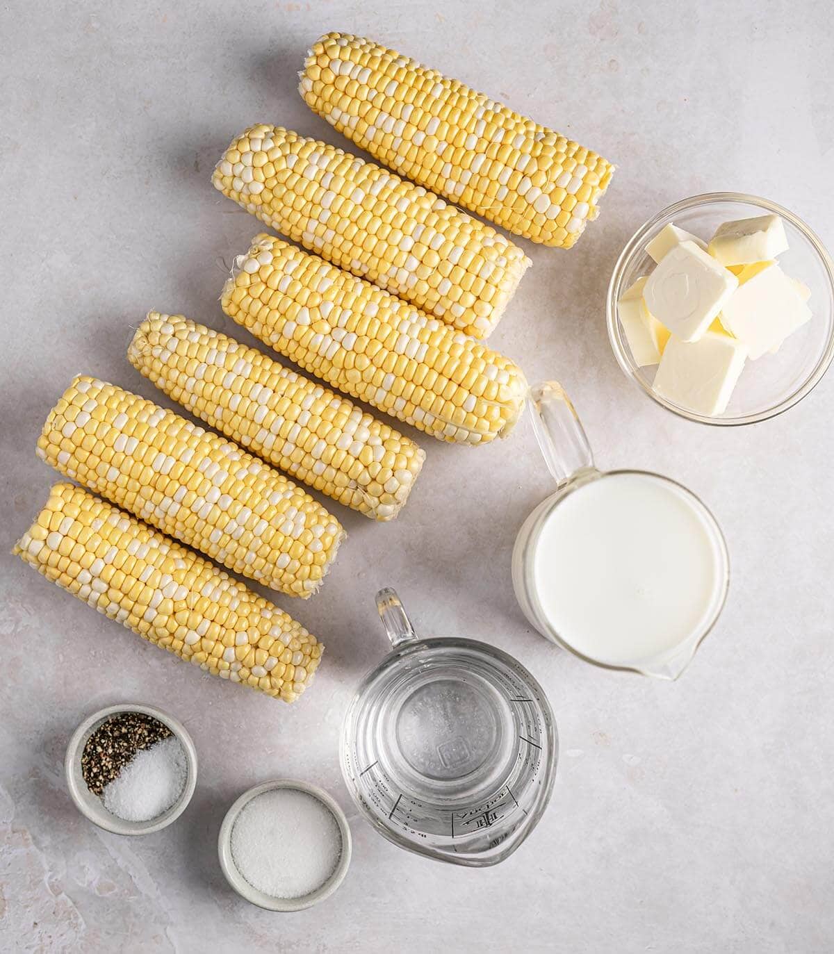 Shucked corn, butter, milk, water, sugar, salt, and pepper laid out on a counter.