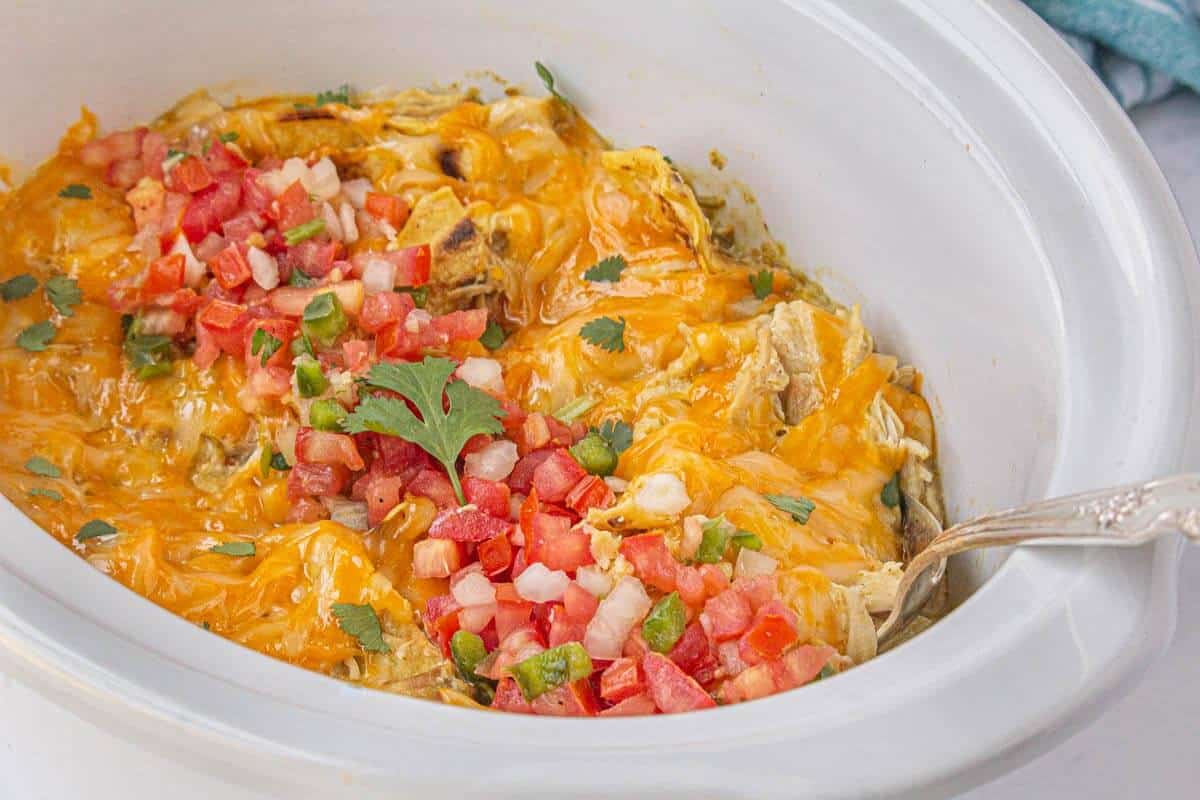Close-up of melted cheese and pico de gallo topping a green chile enchilada casserole in a white slow cooker.