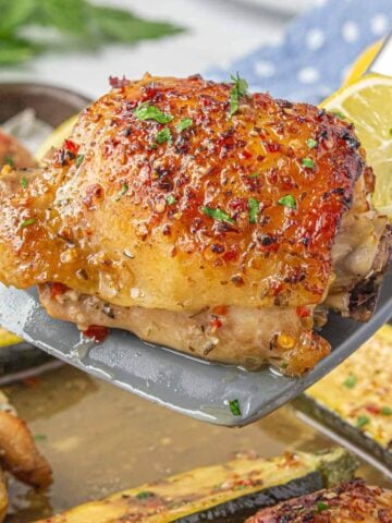 Close-up of a spatula lifting a golden, crispy baked chicken thigh with herbs and red pepper flakes, served with zucchini and lemon.