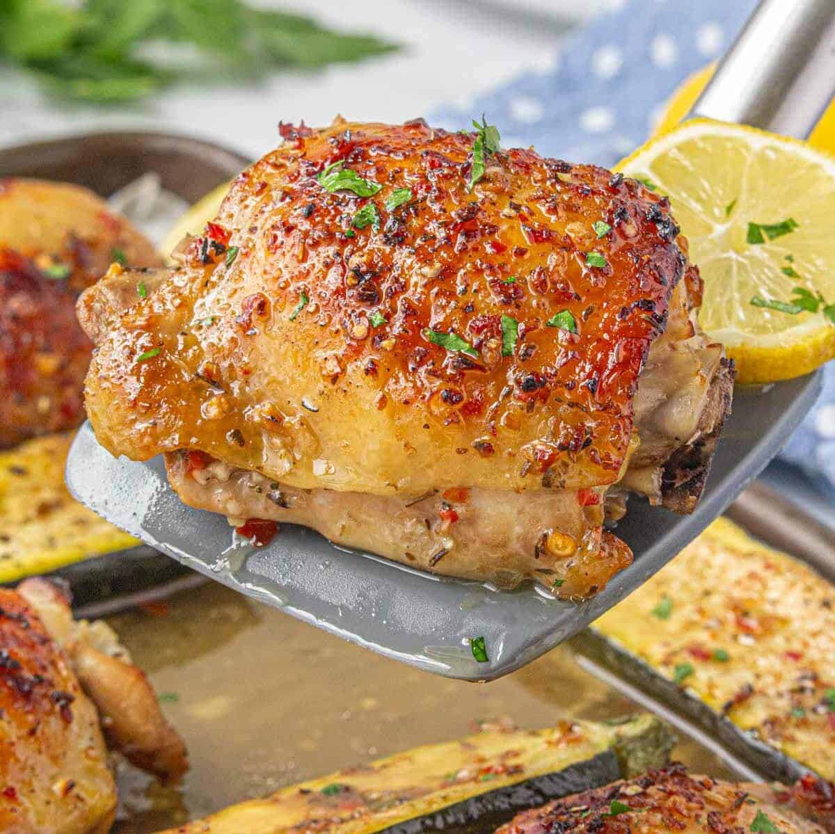 Close-up of a spatula lifting a golden, crispy baked chicken thigh with herbs and red pepper flakes, served with zucchini and lemon.