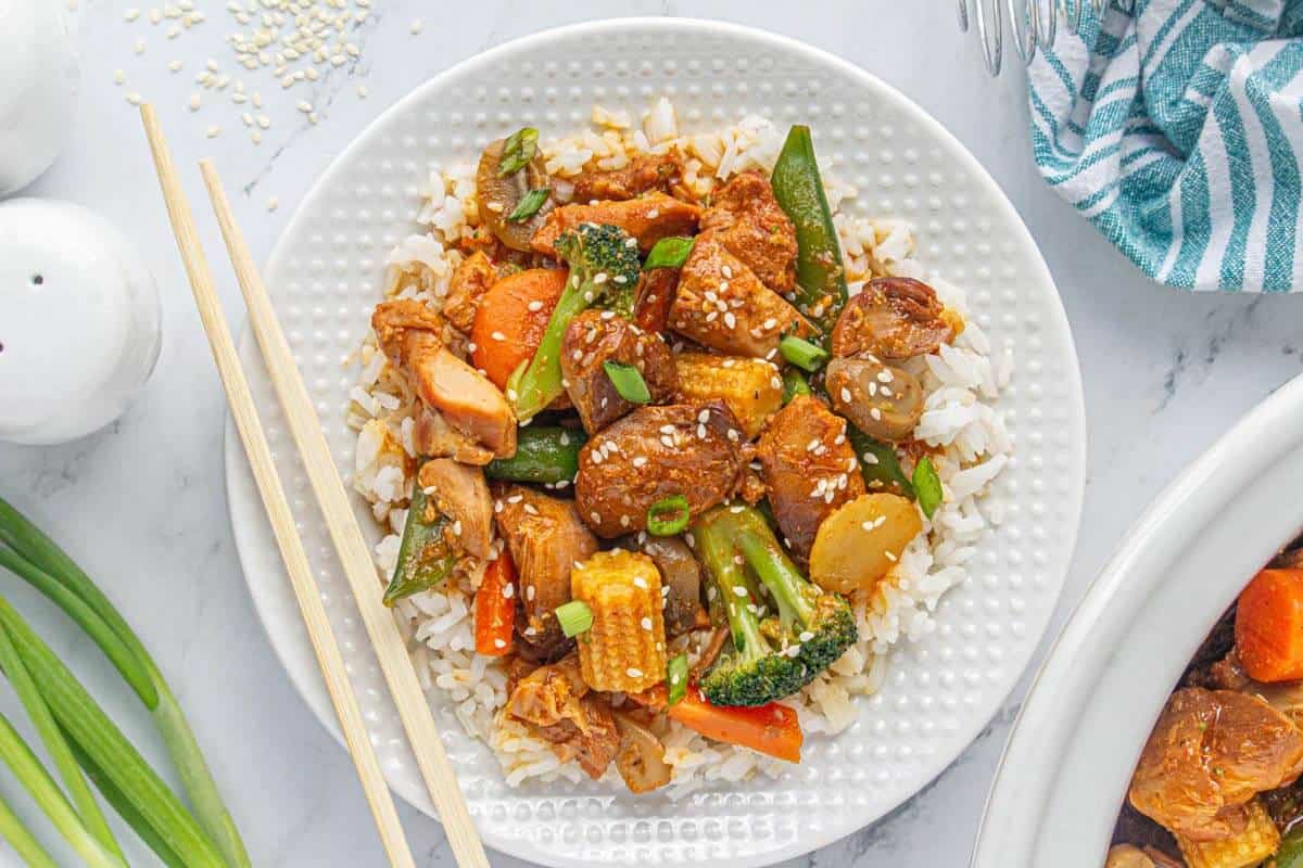 Overhead view of Crockpot chicken stir fry served on a bed of white rice in a white bowl, garnished with sesame seeds and green onions.