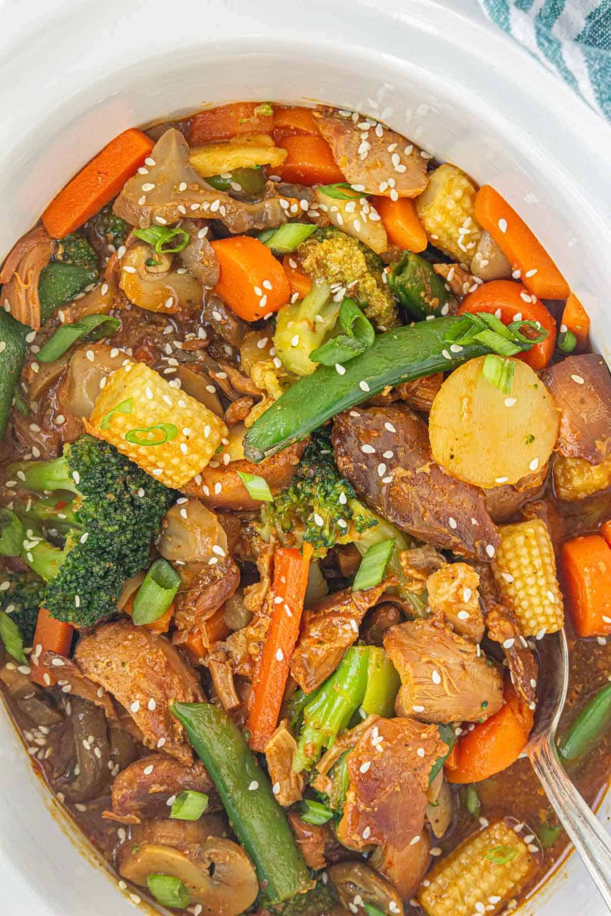 Fully cooked Crockpot chicken stir fry in a white slow cooker, filled with vibrant vegetables and topped with sesame seeds and green onions.