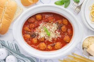 Overhead view of slow cooker filled with saucy meatballs and mushrooms, surrounded by hoagie rolls, dry spaghetti, parmesan, and utensils.