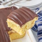 Close-up of a slice of Boston Cream Poke Cake on a serving spatula being lifted from a pan. You can see the fluffy cake, pudding filling, and glossy chocolate top in rich detail. A striped napkin sits alongside.