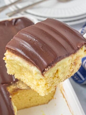 Close-up of a slice of Boston Cream Poke Cake on a serving spatula being lifted from a pan. You can see the fluffy cake, pudding filling, and glossy chocolate top in rich detail. A striped napkin sits alongside.
