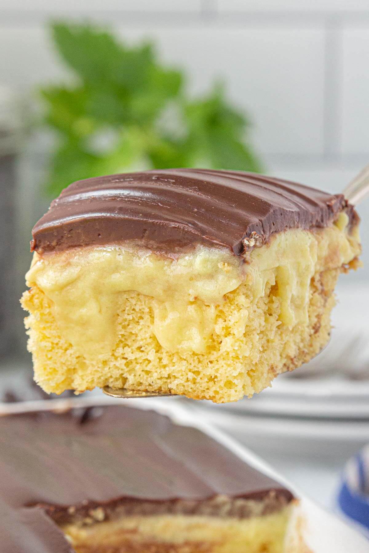 A close-up slice of yellow cake, topped with creamy ganache on a spatula. The rest of the cake is in the background.