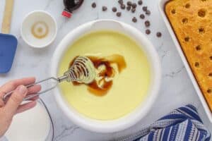 A bowl of yellow pudding mixture with vanilla extract being stirred in using a metal whisk. A baked yellow cake with holes poked in the top sits nearby.