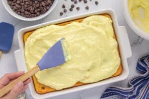A hand spreads creamy vanilla pudding mixture over a yellow poke cake using a blue silicone spatula. A bowl of chocolate chips is visible nearby.