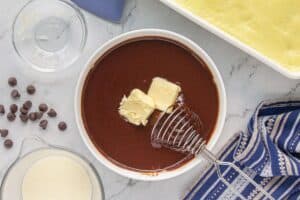 A bowl of melted chocolate ganache with chunks of butter added on top. A whisk is partially submerged in the ganache, ready to stir.