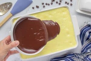 A hand holds a white bowl, pouring silky chocolate ganache over a layer of pudding-covered yellow cake in a white baking dish.