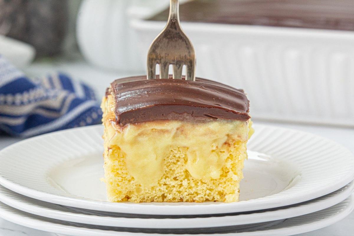 A slice of yellow cake with a creamy pudding filling, topped with a rich ganache on a white plate.