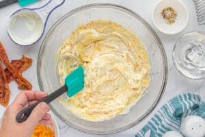 A hand stirs together the creamy mixture of ranch, sour cream, soup, seasoning, and shredded cheese in a large mixing bowl.
