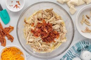 Shredded chicken, diced biscuits and crispy bacon pieces are added to the creamy mixture in a glass mixing bowl.