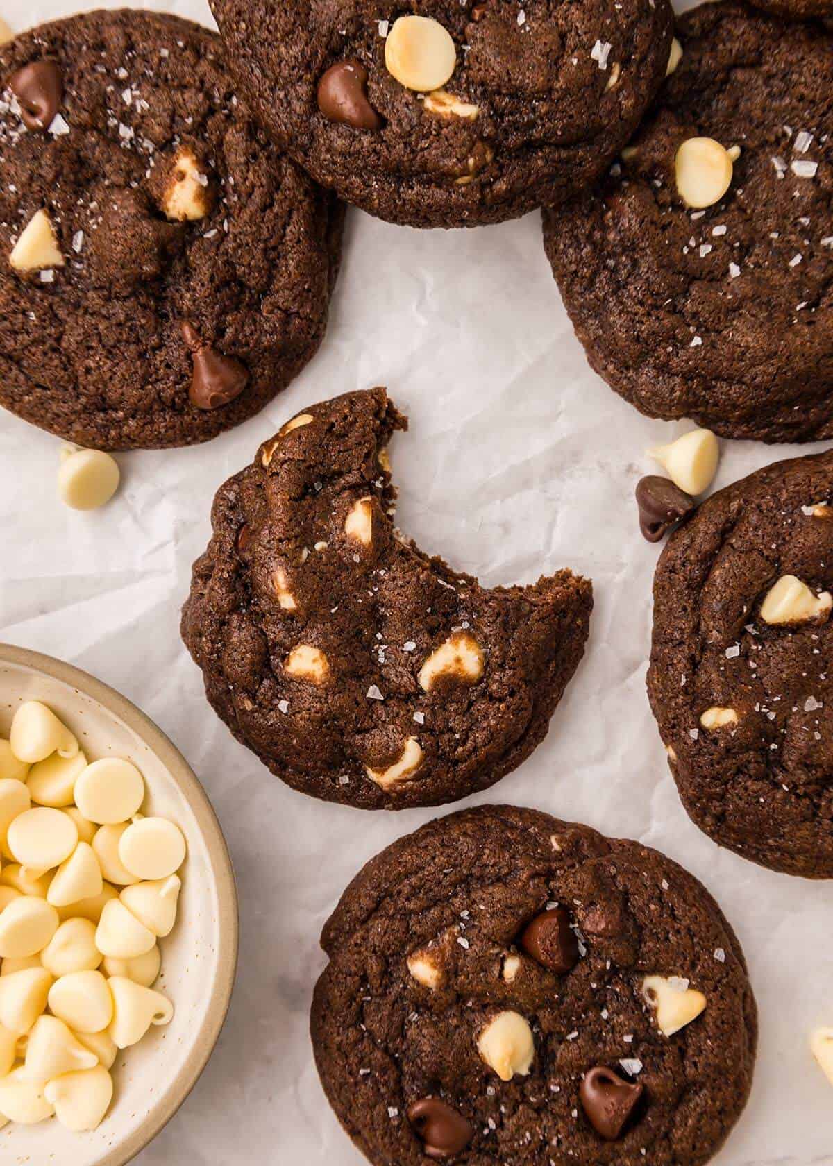 Top-down view of chocolate chocolate cookies with one cookie showing a bite taken out, next to a small bowl of white chocolate chips.