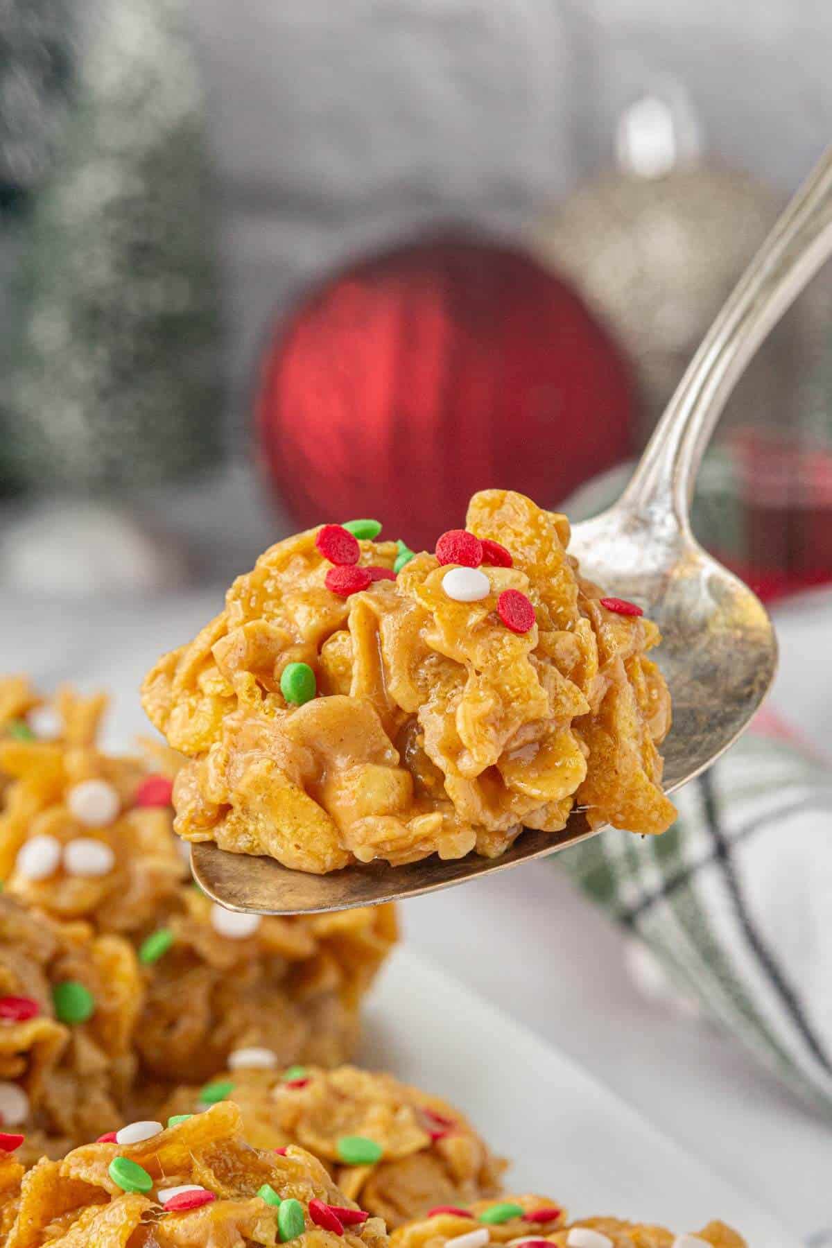 A single peanut butter cornflake cookie topped with red, green, and white sprinkles held up on a silver spoon, with blurred holiday ornaments in the background.