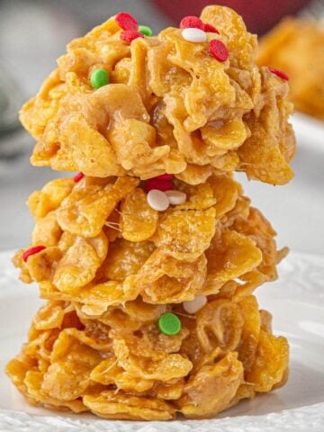 Close-up of three peanut butter cornflake cookies stacked on a white plate, topped with festive red, green, and white sprinkles.