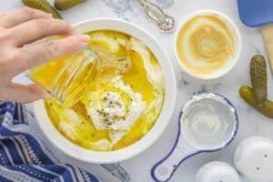 A hand pours pickle juice into a bowl with mayonnaise, mustard, sour cream, and cracked black pepper, prepping the dressing mixture for pasta salad.