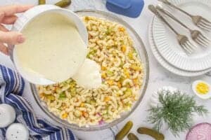 A hand pouring creamy dressing over the assembled pasta salad ingredients in a large glass bowl, surrounded by dill, forks, sliced egg, and pickles.