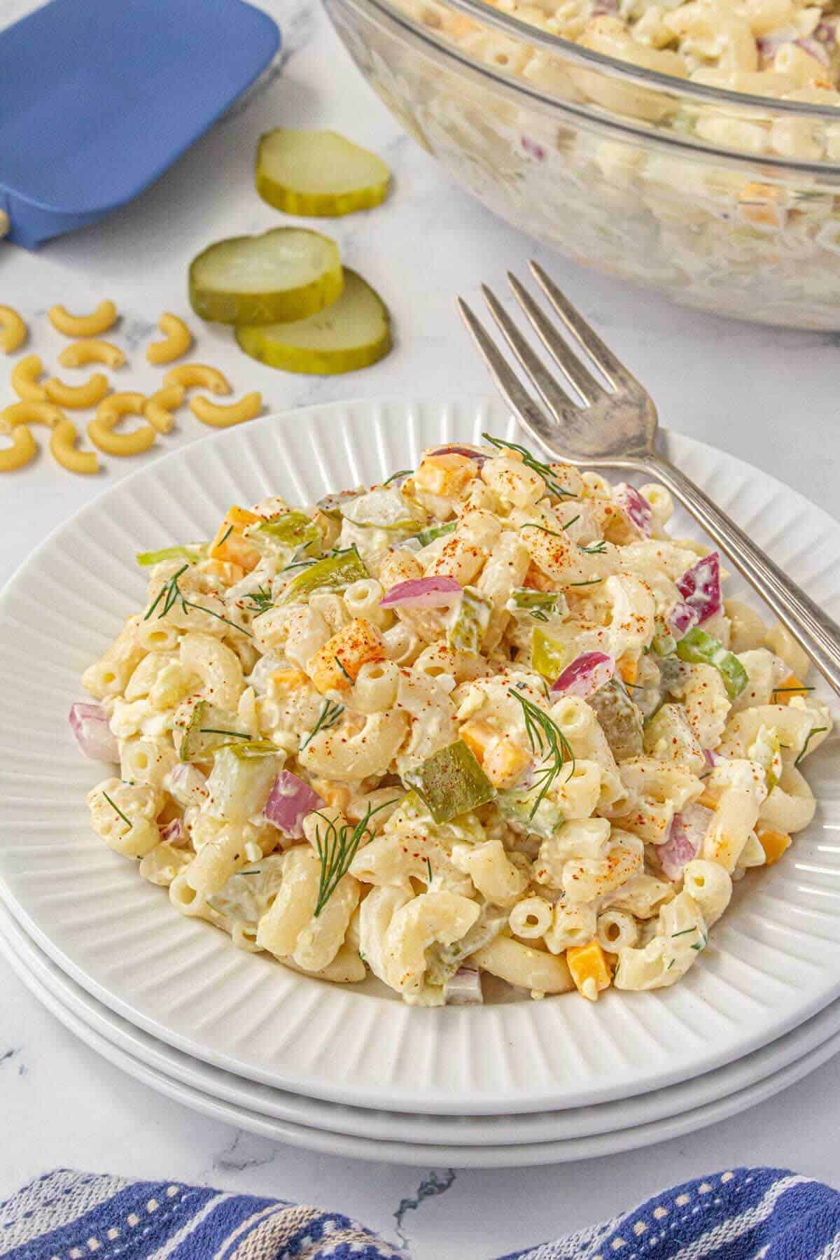 A white plate with a serving of dill pickle pasta salad garnished with fresh dill, surrounded by uncooked macaroni and pickle slices, with a fork resting on the plate.