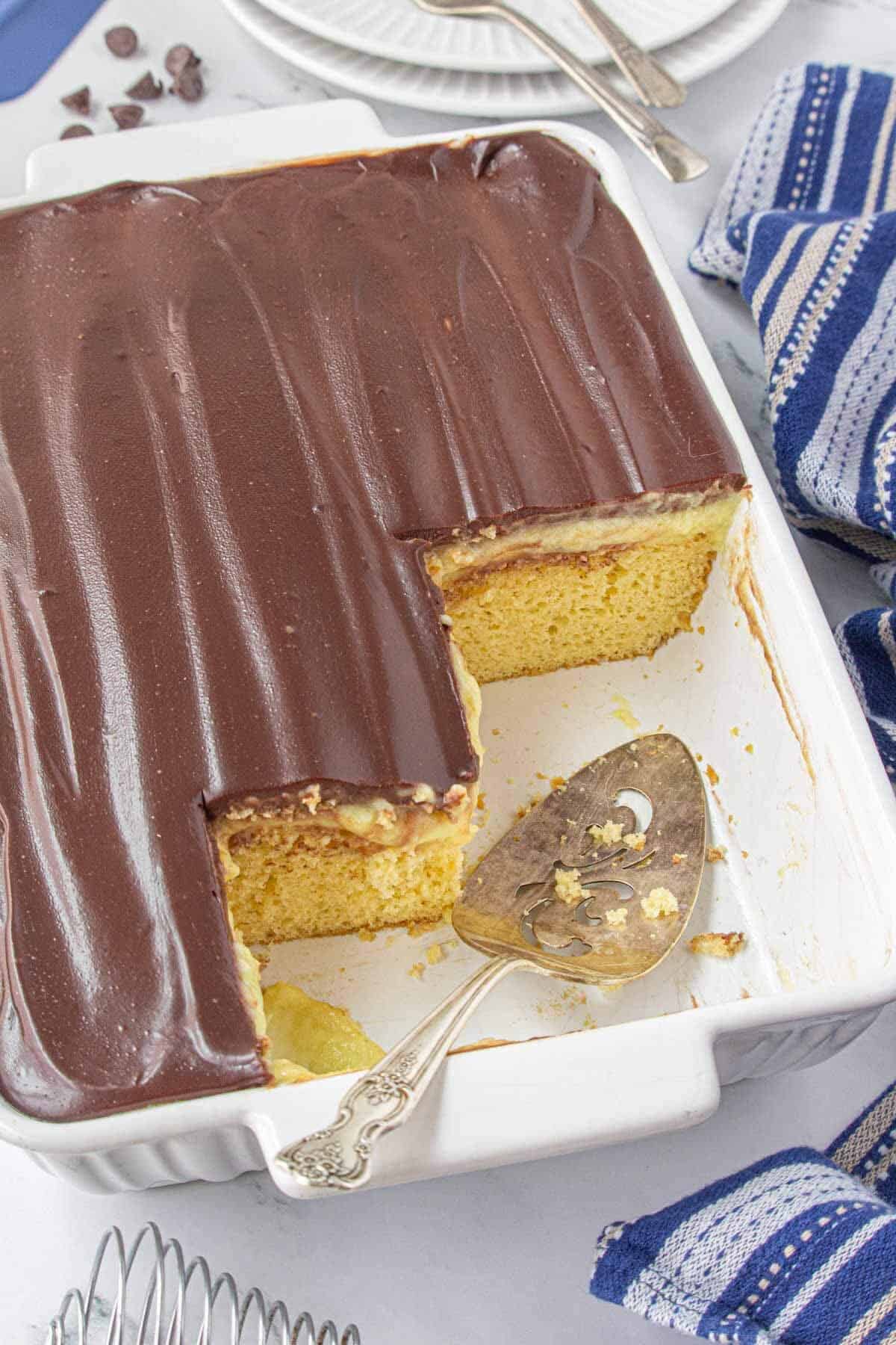 Overhead shot of a rectangular pan filled with Boston Cream Poke Cake, topped with a smooth layer of shiny chocolate ganache. A few slices have been taken out of the corner, showing the soft cake and pudding layers beneath.
