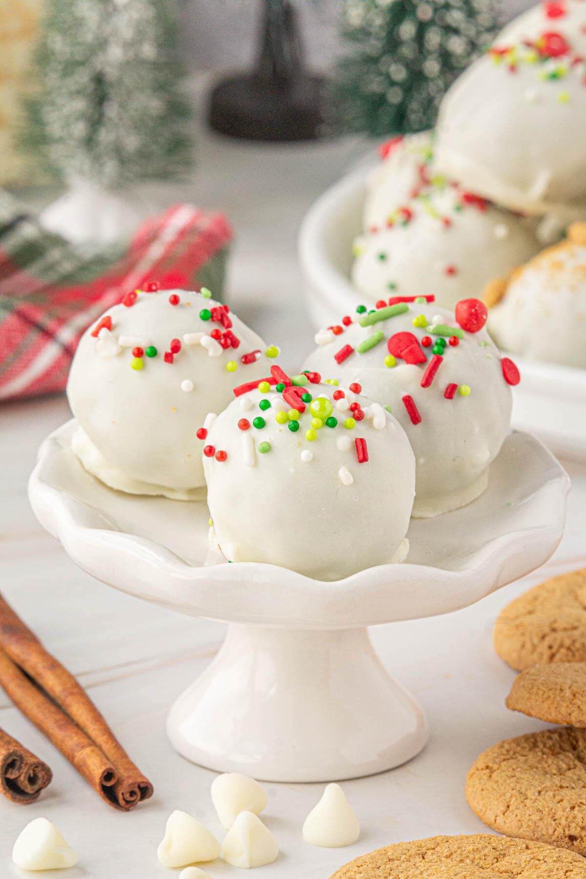 Close-up of three white chocolate-coated gingersnap truffles topped with festive red, green, and white sprinkles, displayed on a white pedestal dish with cinnamon sticks and gingersnap cookies nearby.