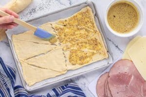 Crescent roll dough rolled out over parchment paper on a sheet pan. A hand is slathering it with mustard spread. Surrounding the dough is slices of ham and cheese.