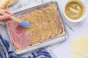 Cheese, ham placed on top of dough and slathered with mustard spread. Additional cheese and sauce sits on the side.