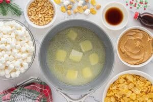 Overhead shot of a saucepan with melting butter, surrounded by ingredients for peanut butter cornflake cookies including marshmallows, chopped peanuts, peanut butter, vanilla, and cornflakes.