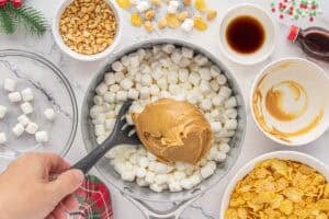 A large spoonful of creamy peanut butter is being added to a pot filled with mini marshmallows. Surrounding bowls hold chopped peanuts, cornflakes, and vanilla extract.