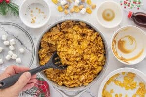 Melted peanut butter and marshmallow mixture stirred together with cornflakes in a saucepan, ready to be shaped into cookies. Empty bowls and extra ingredients sit nearby.