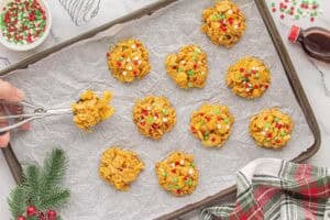 Peanut butter cornflake cookies being portioned onto a parchment-lined baking sheet using a cookie scoop. Each cookie is topped with festive red, green, and white sprinkles.