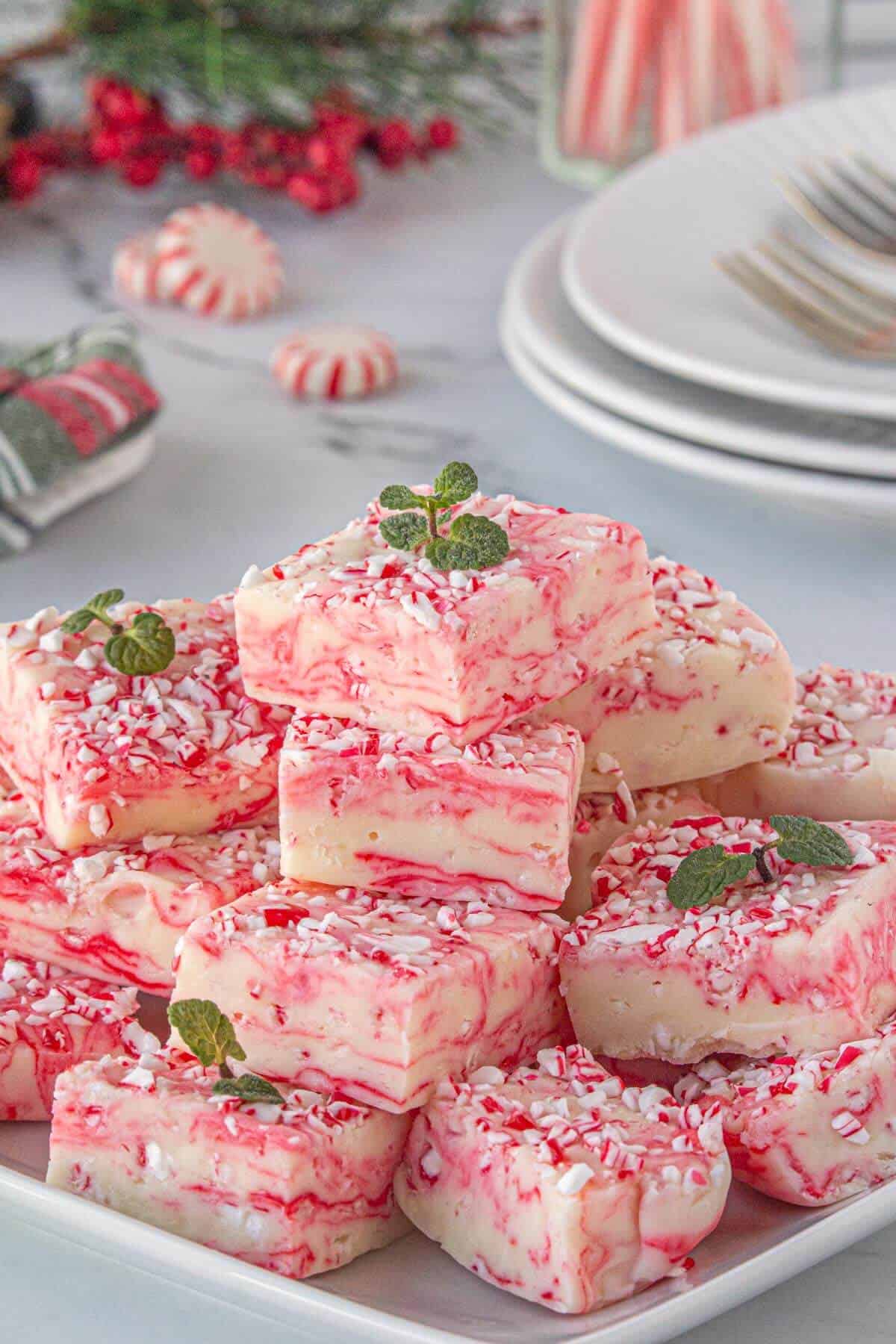 A festive plate stacked with red and white swirled peppermint fudge squares, topped with crushed peppermint and mint leaves, with holiday décor in the background.