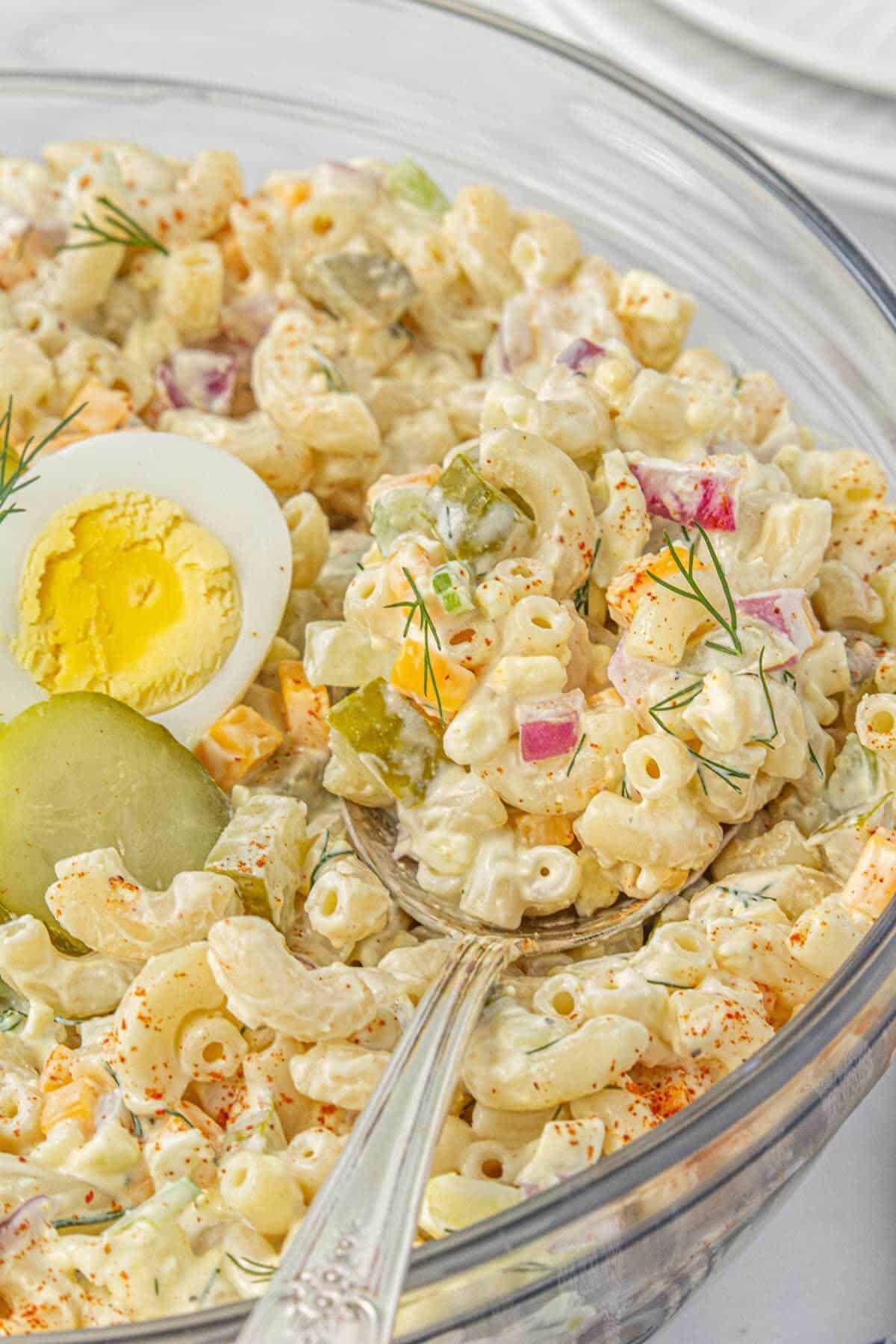 A glass serving bowl filled with dill pickle pasta salad, garnished with sliced pickles, halved boiled eggs, and a sprinkle of paprika and fresh dill.