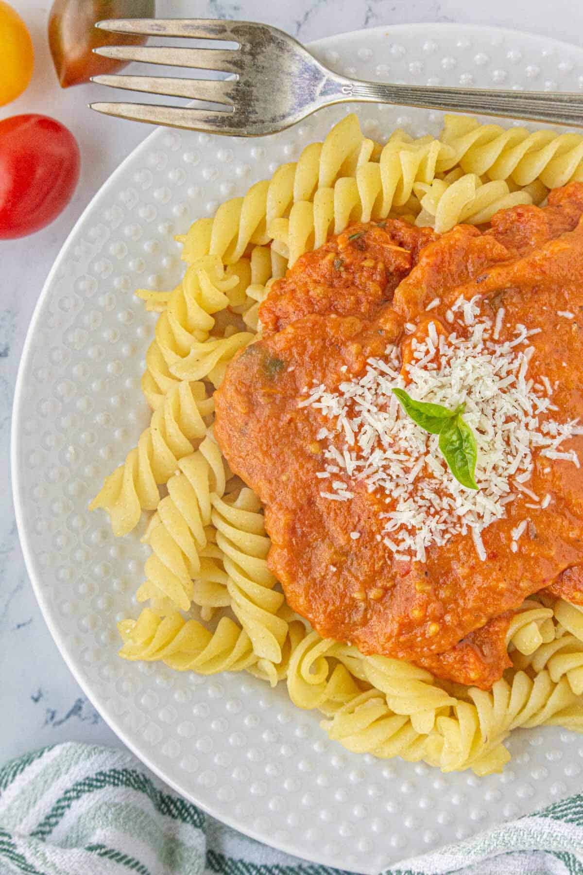 A white plate filled with rotini pasta, topped with tomato garlic pasta sauce, sprinkled with parmesan cheese and fresh basil.