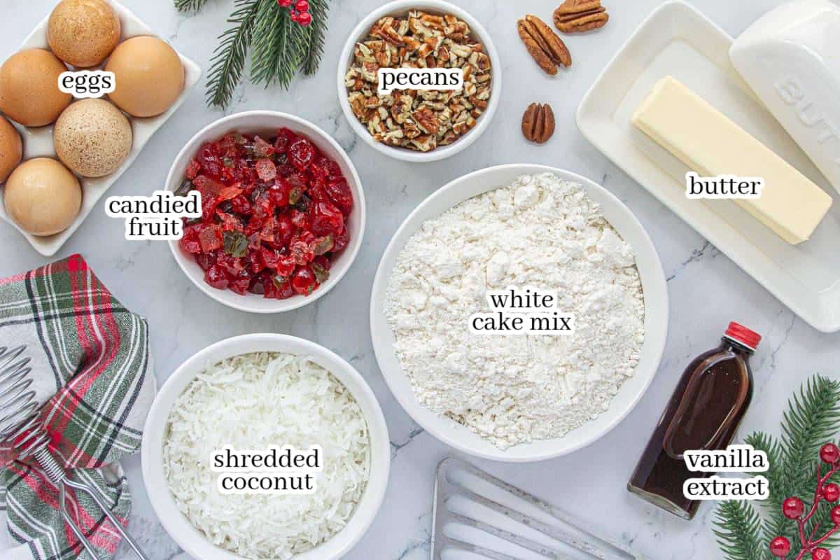 Labeled ingredients for cookie recipe arranged on a countertop, including eggs, candied fruit, pecans, white cake mix, shredded coconut, butter, and vanilla extract. Holiday greenery and a plaid towel add festive touches.