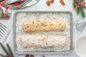Two dough logs rolled in shredded coconut on a baking sheet, ready to be chilled. Holiday greenery and baking tools surround the scene.