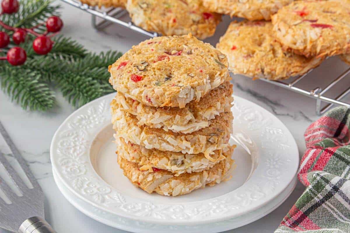 Stack of baked cookies with coconut edges and colorful cherry pieces on a white plate, with more cookies cooling in the background and festive decor nearby.