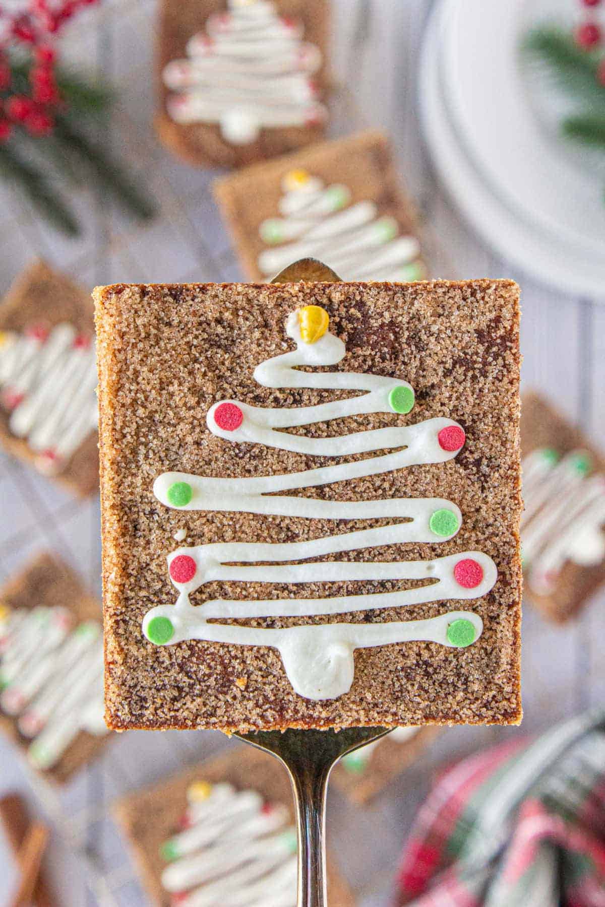 A close-up of a snickerdoodle cookie bar, decorated with white frosting in the shape of a Christmas tree, decorated with colorful sprinkles. It is being held by a spatula, with more cookie bars in the background.