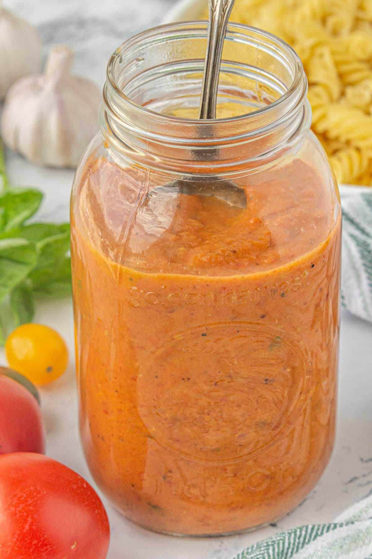 A glass tomato jar filled with tomato sauce with a spoon inside. Surrounded by tomatoes, garlic and pasta.