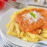 Rotini pasta smothered in roasted tomato and garlic pasta sauce on a white plate topped with Parmesan cheese and fresh basil. With tomatoes and garlic in the background.