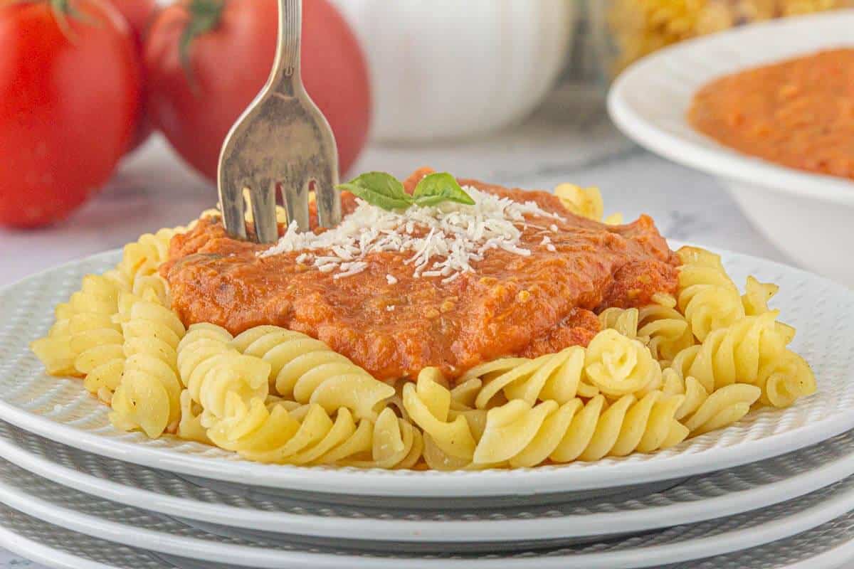 A fork poised over a plate filled with rotini pasta topped with tomato garlic sauce sprinkled with parmesan cheese and fresh basil.