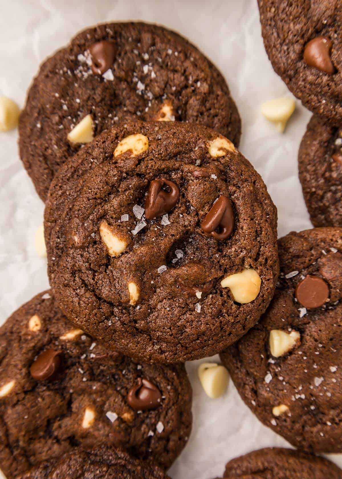 Overhead shot of several triple chocolate cookies with white and semi-sweet chocolate chips and a dusting of sea salt on parchment paper.