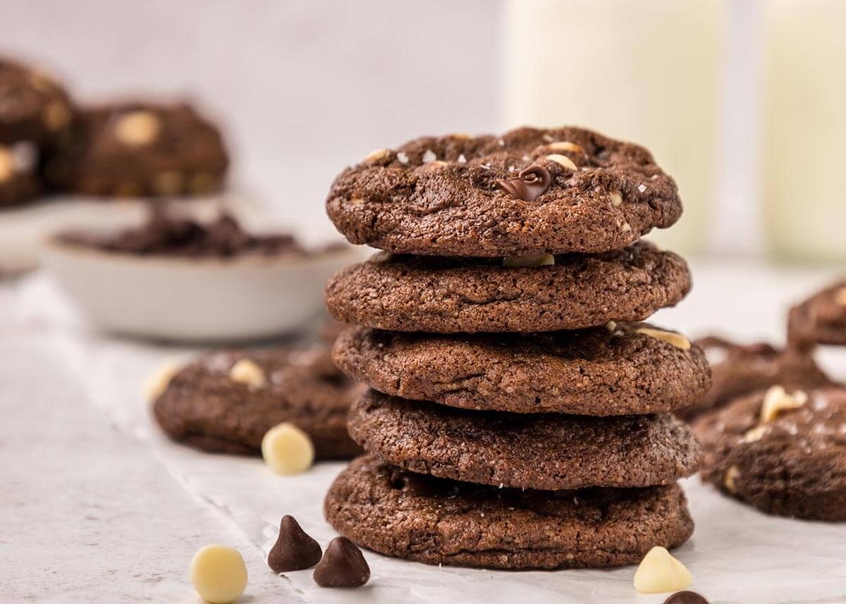 Stack of five thick and chewy triple chocolate cookies with chocolate chips scattered around and a glass of milk in the background.