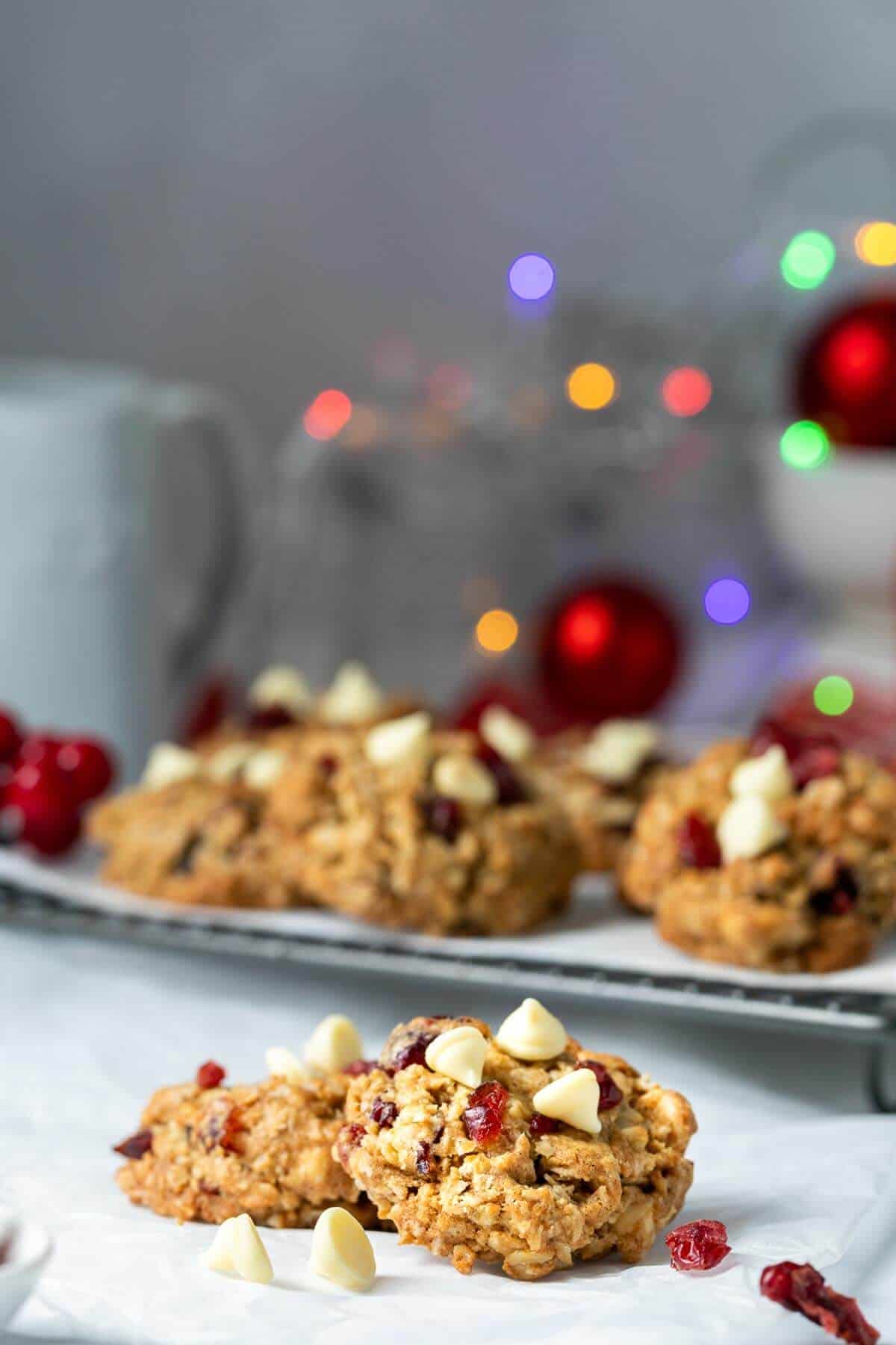 Festive holiday setup featuring cranberry white chocolate oatmeal cookies on a wire rack, with Christmas lights and red ornaments softly glowing in the background.
