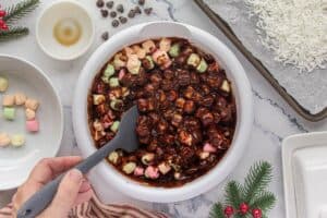 Multi colored mini marshmallows being stirred into melted chocolate with a spatula.