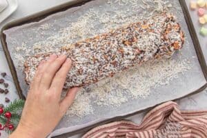 Shredded coconut being pressed into roll of chocolate candy on a sheet of parchment paper.